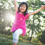 Little girl with healthy baby teeth in a pink dress smiles while playing outside